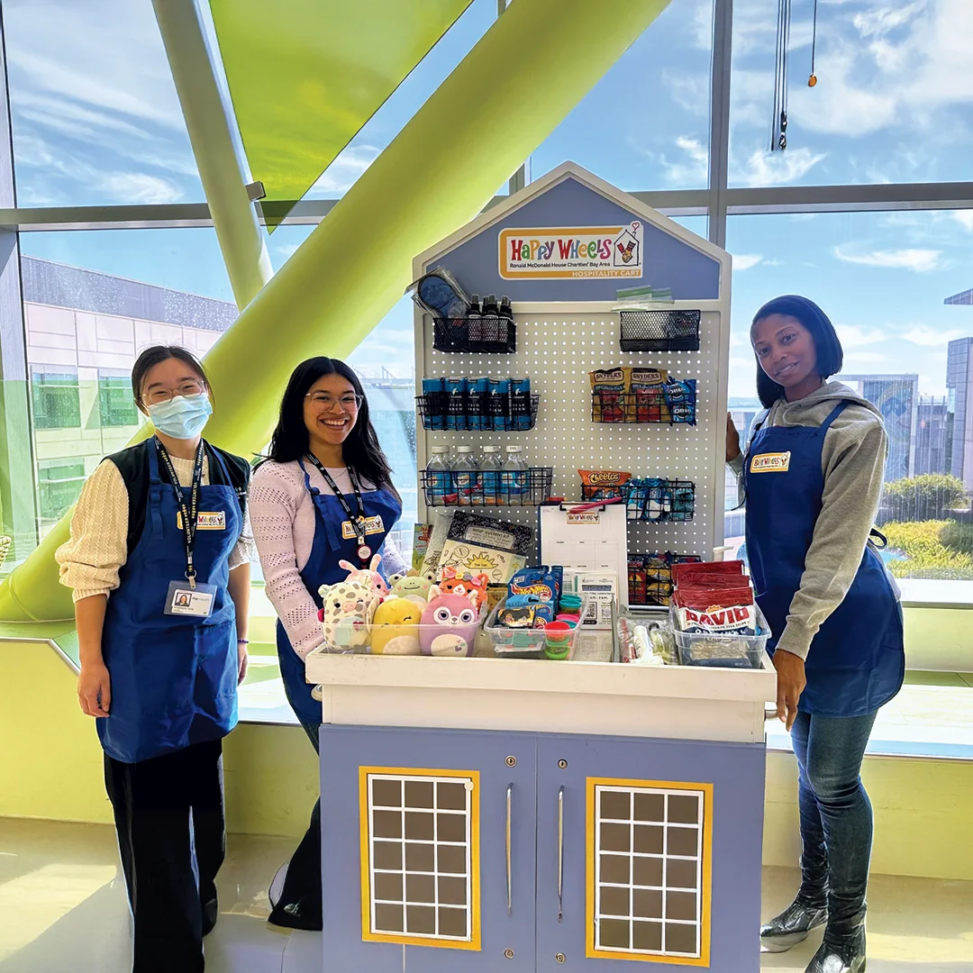RMHC Bay Area team members stand beside a fully stocked Happy Wheels cart filled with snacks, toys, and supplies at UCSF Mission Bay.