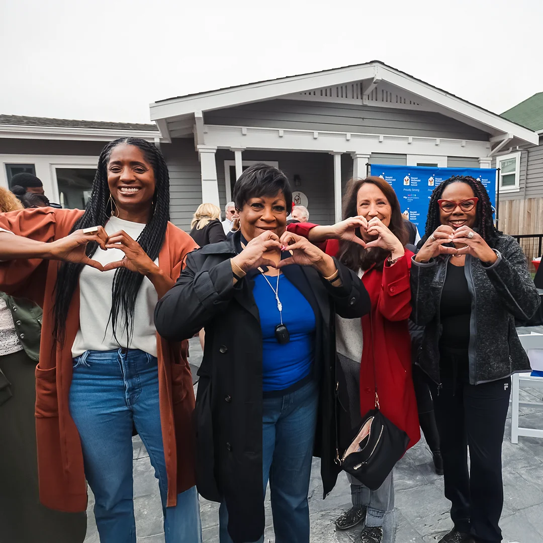 Four smiling women stand together in front of an RMHC Bay Area house, forming heart shapes with their hands in a show of support.